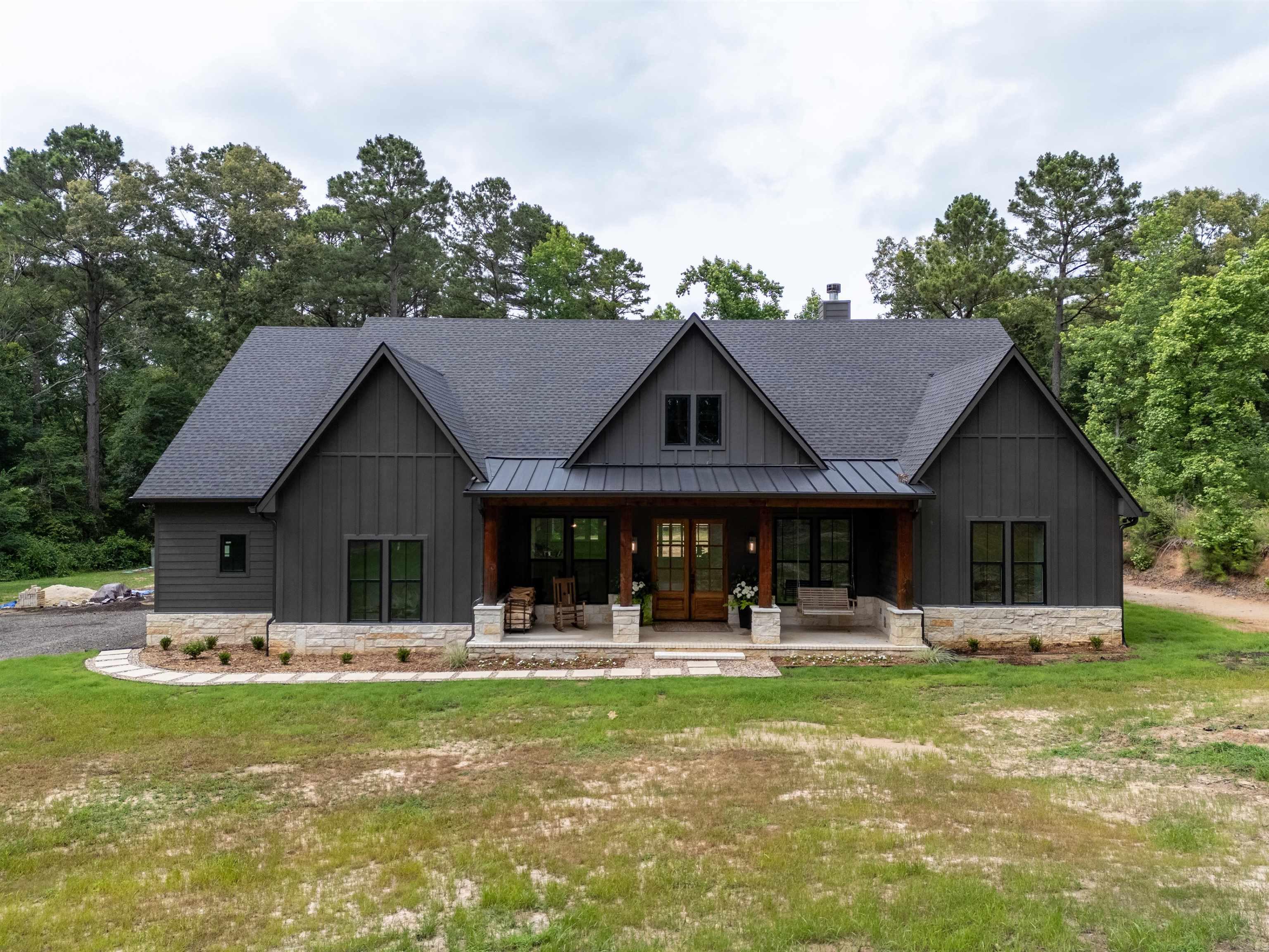 Image 1: Modern farmhouse style residence featuring dark vertical siding, a stone foundation, a covered front porch with substantial wood columns, a dark shingled roof, and multiple large windows, Front Of Structure