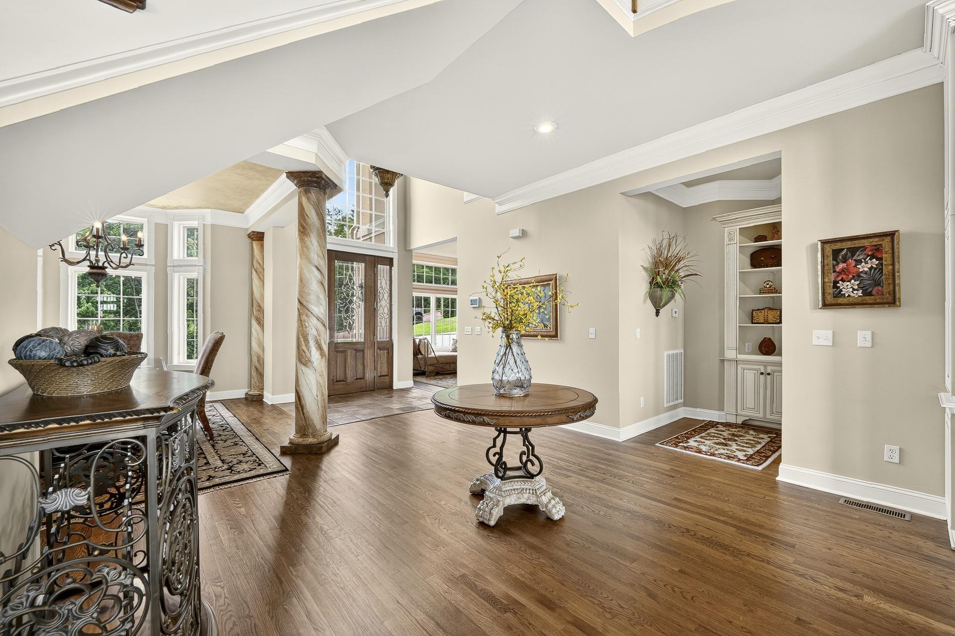 Image 3: Spacious foyer featuring rich wood-finish flooring, decorative architectural columns, a double-door entry with transom windows, a built-in display cabinet, and extensive crown molding