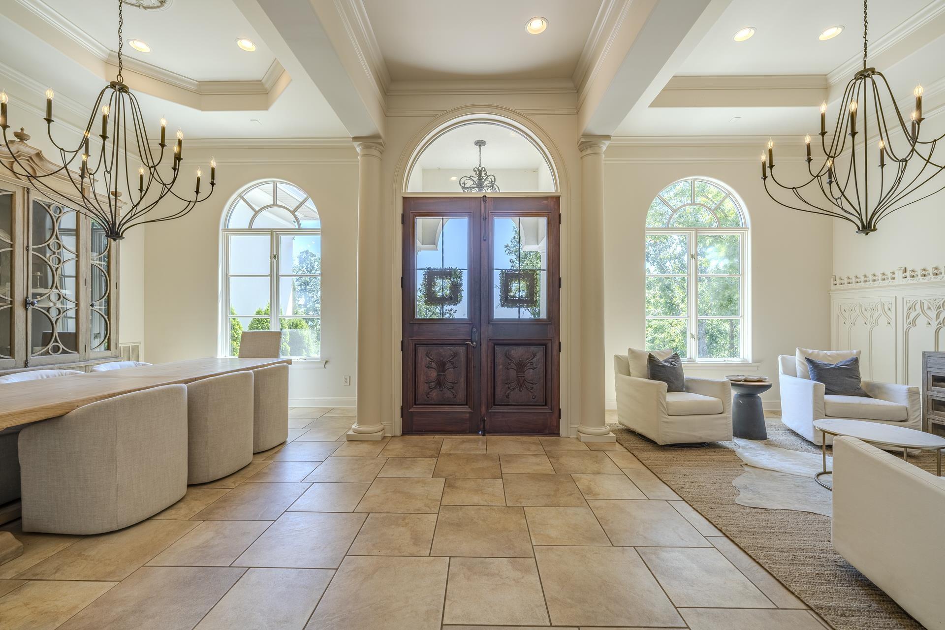 Image 3: Grand entry foyer featuring an arched dark wood double door, coffered ceiling, two large iron chandeliers, and large format tile flooring