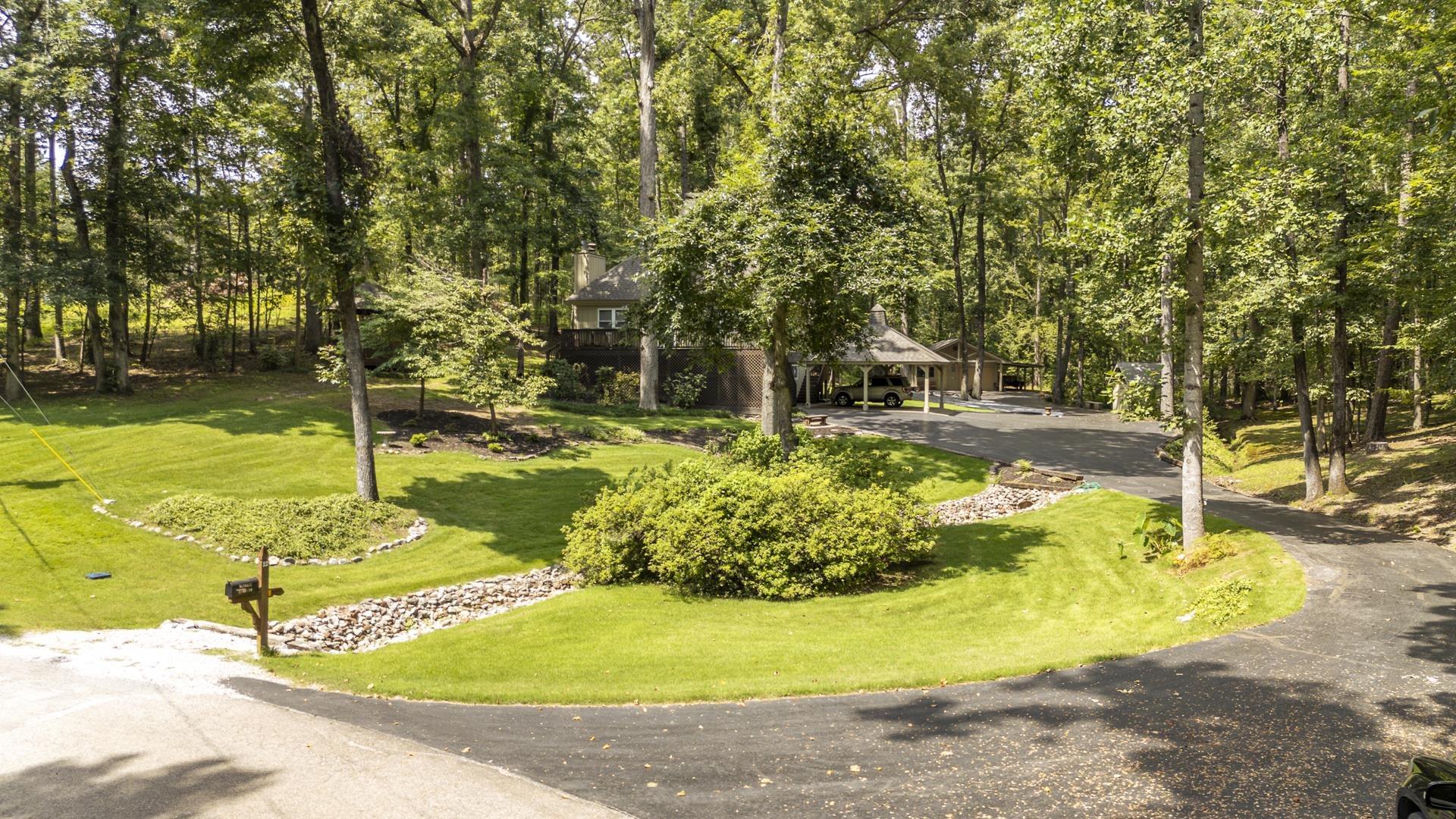 Image 1: Mature wooded setting with expansive manicured lawn, stone-bordered garden beds, and asphalt driveway