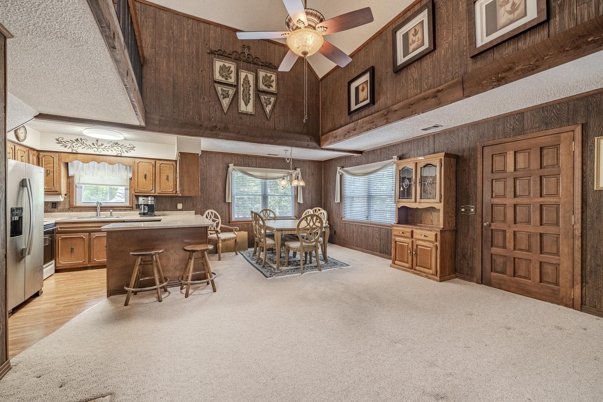 Image 4: Vaulted ceiling with exposed beam, wood paneling, light wood cabinetry, kitchen island with breakfast bar, and stainless steel refrigerator