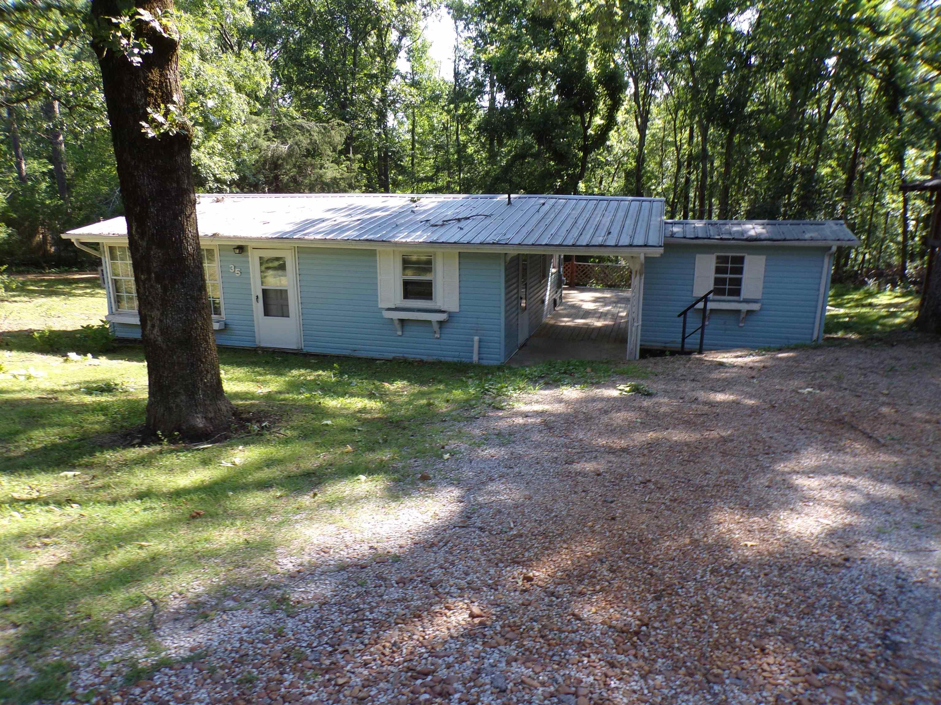 Image 3: Light blue siding exterior featuring a white door, white-framed windows with decorative shutters, and a covered carport