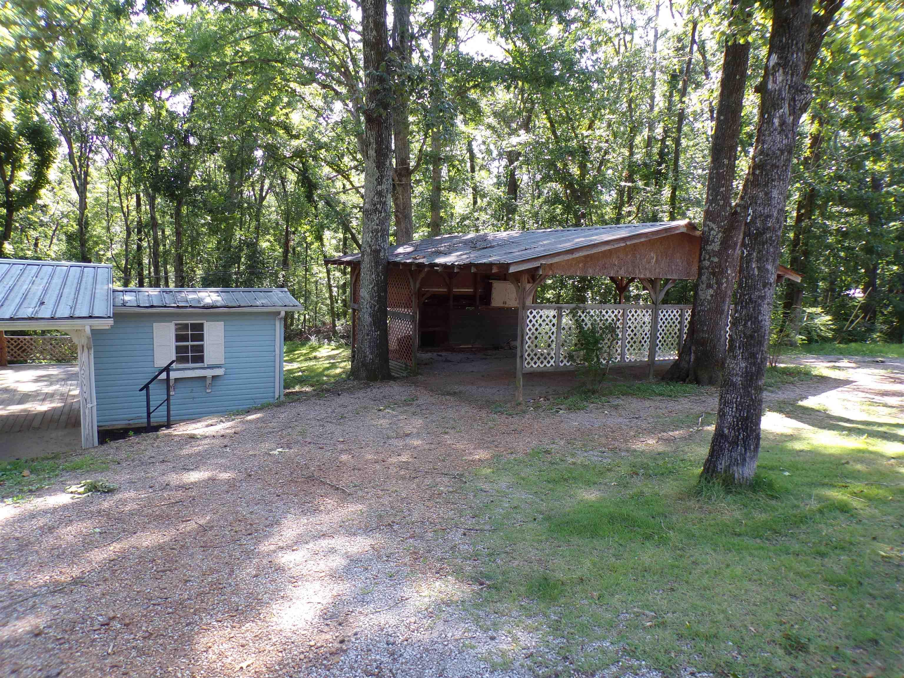 Image 4: Gravel driveway with tree-shaded covered structure, blue siding shed with white trim window, and carport