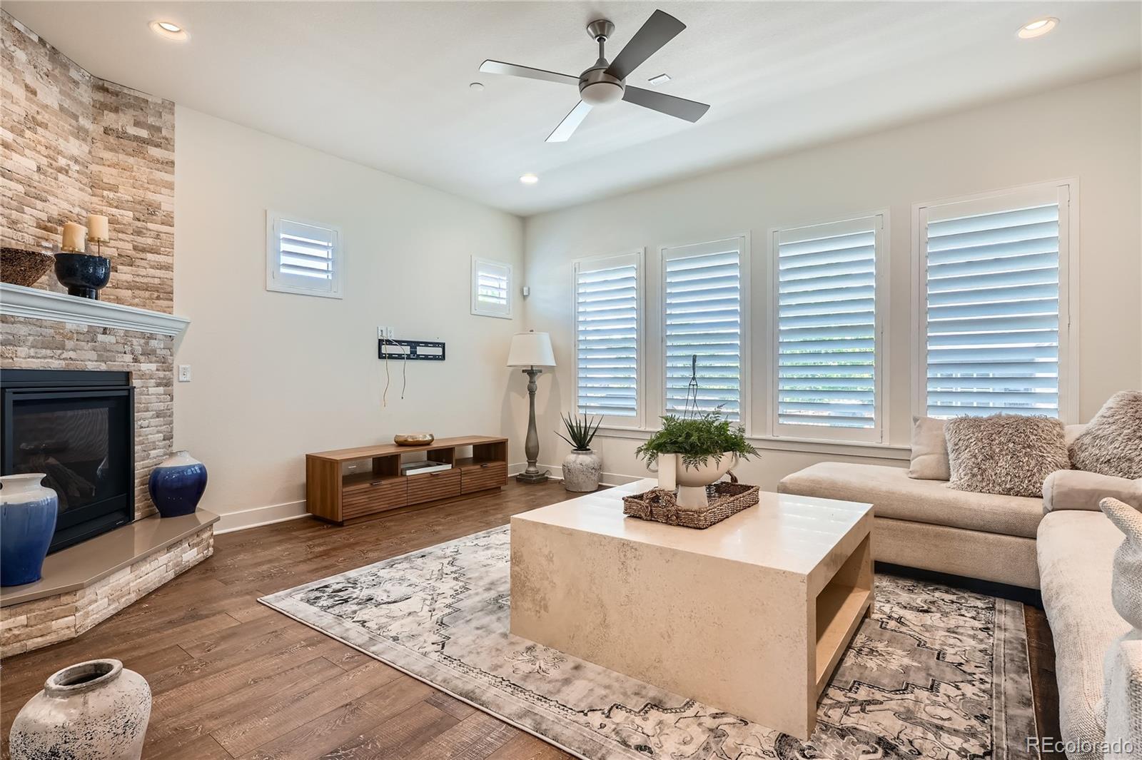 Image 4: 18x16 sq ft Family Room with stone-wall fireplace, wood floors, plantation shutters, and ceiling fan with a light.