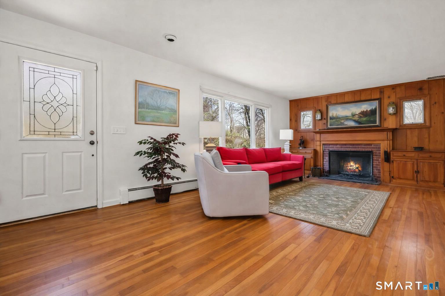 Image 2: Warm and inviting living room featuring beautiful hardwood floors, a classic brick fireplace framed by rich wood paneling, and a large window that bring in wonderful natural light. A perfect space for relaxing evenings by the fire.