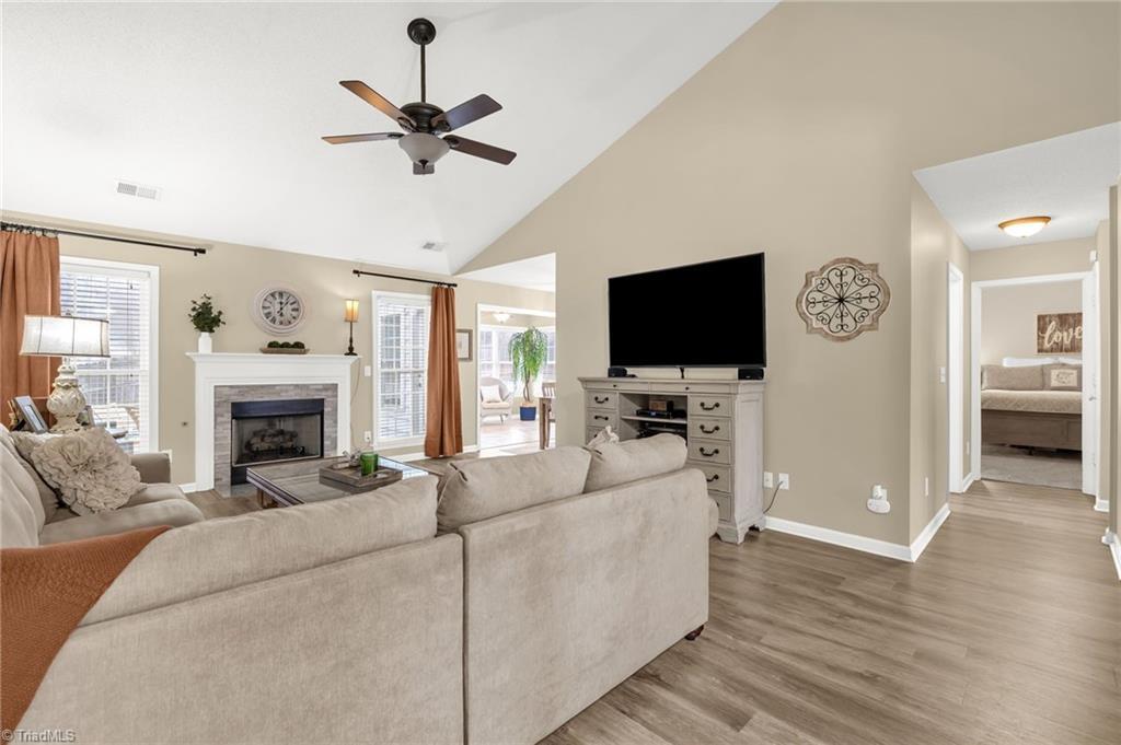 Image 2: A bright, inviting living room with vaulted ceilings, warm neutral tones, and a cozy fireplace as the focal point. Large windows bring in natural light, while the open layout flows seamlessly into the sunroom and nearby bedroom hallway.